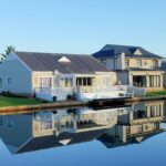 Luxury modern homes reflecting on a peaceful, clear lake during daylight, showcasing architectural beauty.